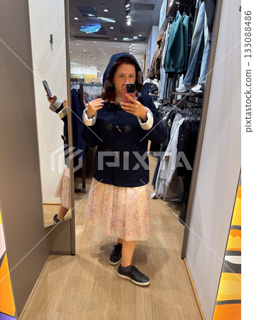 Woman trying on hooded navy duffle coat with toggle closure in fitting room. Warm casual style symbolizing coziness, simplicity, and seasonal charm. 133088486