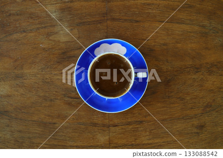 Close-up and top view of hot black coffee in blue coffee cup and roasted Thai coffee beans on wooden background. Close-up and top view of hot black coffee in blue coffee cup and roasted Thai coffee beans on wooden background. 133088542