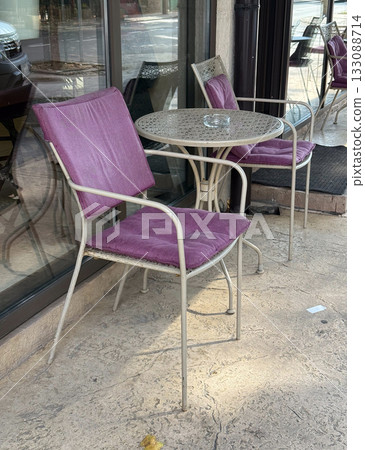 Outdoor cafe chairs with purple cushions near glass window. Urban leisure, street relaxation, and cozy city atmosphere 133088714