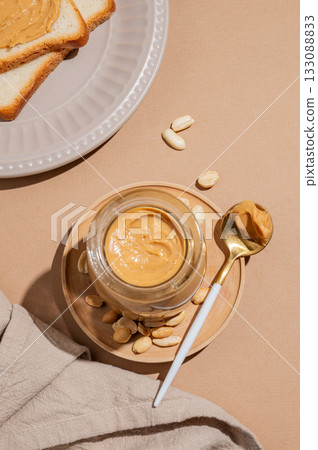 Peanut butter in a glass jar and spoon on a beige background with toast Peanut butter in a glass jar and spoon on a beige background with toast 133088833