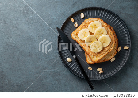 Peanut butter toast and banana on a black plate on a dark background with nuts and knife. 133088839