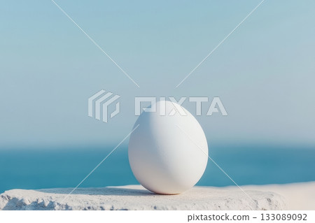 A white egg is placed on a stone surface, showcasing a clear blue sky and ocean in the background during a sunny day 133089092