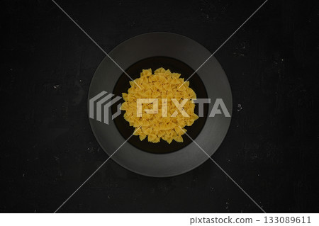 Raw pasta farfalle on plate on black background. Top view of Italian cuisine ingredient. Raw pasta farfalle on plate on black background. Top view of Italian cuisine ingredient. 133089611