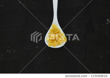 Raw pasta farfalle on wooden spoon on black background. Top view of Italian cuisine ingredient. 133089616