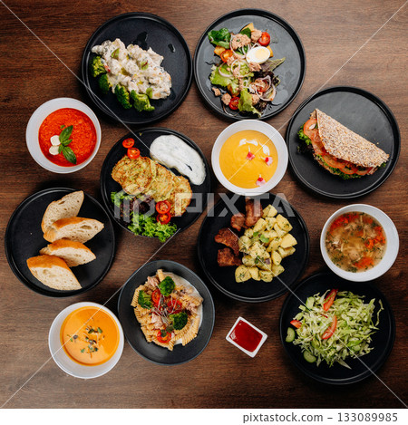 Assorted restaurant dishes flat lay on wooden table with soups salads mains bread and sauces Assorted restaurant dishes flat lay on wooden table with soups salads mains bread and sauces 133089985