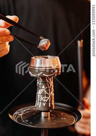 Hand placing hot hookah coal with tongs into metal bowl, warm bar bokeh light 133089991