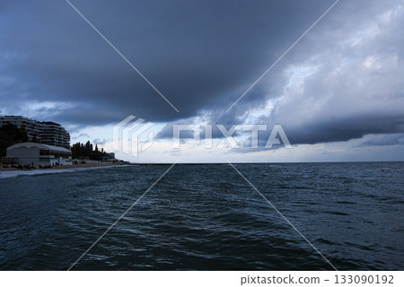 View of the coastal slopes of Odessa with a dark bay and cloudy sky. Dark, cloudy sky looms over a seaside town along the coast. The sea is deep and choppy, stretching towards the horizon.  133090192