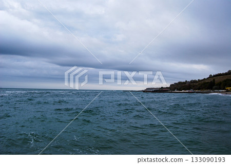 View of the coastal slopes of Odessa with a dark bay and cloudy sky. Dark, cloudy sky looms over a seaside town along the coast. The sea is deep and choppy, stretching towards the horizon.  133090193
