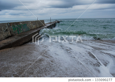 Autumn sea waves lap against a concrete pier. The concrete pier juts into the stormy sea waves under a cloudy sky, creating a gloomy coastal landscape. 133090277