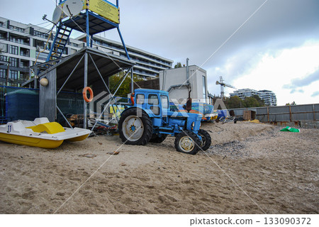 Blue tractor and paddle boat on a sandy beach with lifeguard tower. Blue tractor parked on a sandy beach near a yellow and white paddle boat. Blue tractor and paddle boat on a sandy beach with lifeguard tower. Blue tractor parked on a sandy beach near a yellow and white paddle boat. 133090372