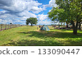 Guests set up tents on a grassy field under a blue sky with clouds near a wooden fence 133090447