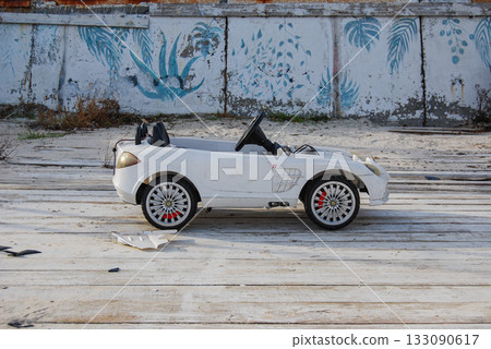 An abandoned white toy car against a concrete wall with leaf patterns. A small white toy convertible car sits on a worn wooden deck on a sandy beach. An abandoned white toy car against a concrete wall with leaf patterns. A small white toy convertible car sits on a worn wooden deck on a sandy beach. 133090617