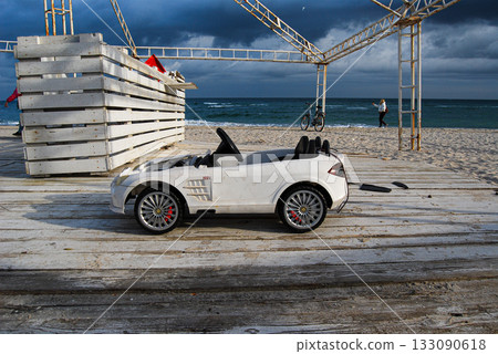 White toy car on a beach boardwalk under a cloudy sky. A white toy car sits on a wooden beach boardwalk under a cloudy sky. White toy car on a beach boardwalk under a cloudy sky. A white toy car sits on a wooden beach boardwalk under a cloudy sky. 133090618