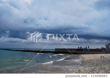 Dramatic coastal view under dark clouds. The turbulent waves of the Black Sea crash onto the sandy beach of Odessa under a sky full of dark, dramatic clouds. 133090663