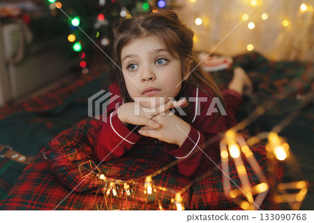 Dreamy child girl in red pajamas lies on festive bed with garland next to Christmas tree waiting for holiday at home 133090768