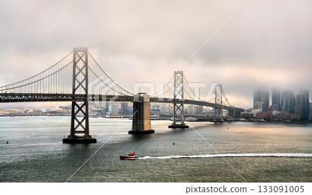San Francisco-Oakland Bay Bridge emerges from thick fog, with the city skyline partially obscured. A small red boat travels across the bay in the foreground. This image has a moody, atmospheric feel 133091005