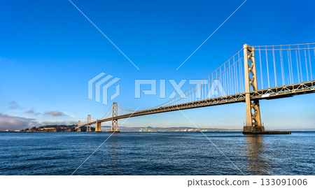 A wide, low-angle view of the San Francisco-Oakland Bay Bridge from the water. The structure spans the bay under a clear, vibrant blue sky. This image represents travel, engineering, and Bay Area 133091006