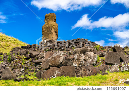 Low-angle view of the Moai statue on Ahu Ature at Anakena Beach, Easter Island. The lone stone ahu sits on a grassy hill under a blue sky with clouds 133091007