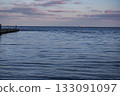 Tranquil Seascape with Concrete Pier at Dusk. Concrete pier extends into a calm seascape at dusk. The water is a deep blue, reflecting the pink and purple hues of the evening sky. 133091097