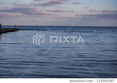 Tranquil Seascape with Concrete Pier at Dusk. Concrete pier extends into a calm seascape at dusk. The water is a deep blue, reflecting the pink and purple hues of the evening sky. 133091097
