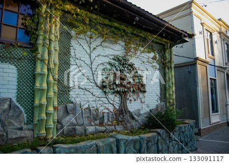 Ivy-covered wall with faux tree and bamboo accents. An exterior wall features an ivy-covered surface with a faux tree design, using metal leaves and branches for a realistic effect. Ivy-covered wall with faux tree and bamboo accents. An exterior wall features an ivy-covered surface with a faux tree design, using metal leaves and branches for a realistic effect. 133091117