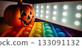 Halloween glow jack-o-lantern with rainbow pop-it and led lights. Jack-o-lantern with carved face, casting an orange glow. Beside it, a rainbow-colored pop-it toy with bubble-textured surface. 133091132