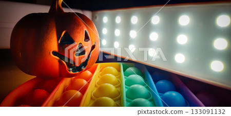 Halloween glow jack-o-lantern with rainbow pop-it and led lights. Jack-o-lantern with carved face, casting an orange glow. Beside it, a rainbow-colored pop-it toy with bubble-textured surface. Halloween glow jack-o-lantern with rainbow pop-it and led lights. Jack-o-lantern with carved face, casting an orange glow. Beside it, a rainbow-colored pop-it toy with bubble-textured surface. 133091132
