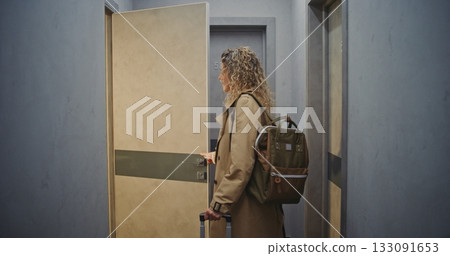 Confident Female Guest in Coat And Sneakers Walks Corridor With Suitcase, Finds Room, Steps Inside 133091653