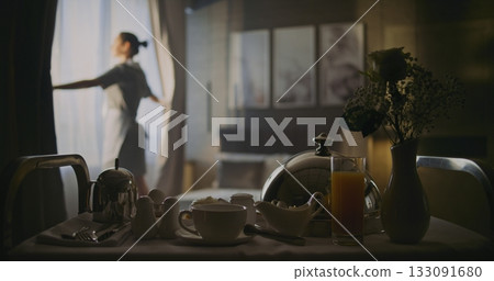 Professional Hotel Maid Opens Curtains on Windows and Makes Fresh, Neat Bed in Elegant Room 133091680