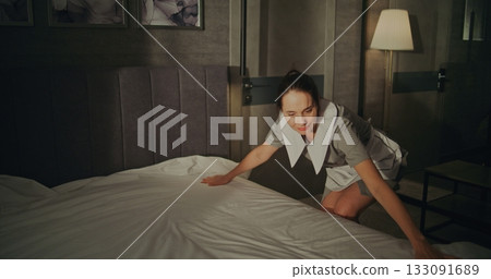 Cheerful Female Maid in Professional Hotel Uniform Smooths Perfectly Fresh Bedding on Large Bed 133091689