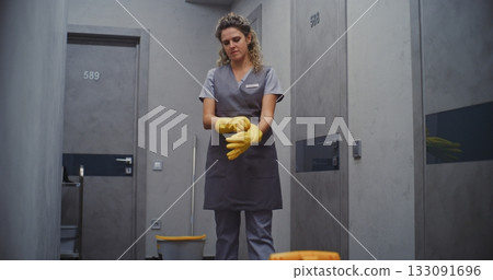 Female Hotel Maid Adjusts Yellow Rubber Gloves in Hallway, Getting Ready to Clean Guest Room Female Hotel Maid Adjusts Yellow Rubber Gloves in Hallway, Getting Ready to Clean Guest Room 133091696