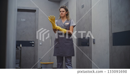 Female Hotel Maid Adjusts Yellow Rubber Gloves in Hallway, Getting Ready to Clean Guest Room Female Hotel Maid Adjusts Yellow Rubber Gloves in Hallway, Getting Ready to Clean Guest Room 133091698