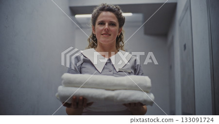 Smiling Hotel Maid in Classic Uniform Carries Fresh White Towels in Hallway, Ready to Prepare Room 133091724