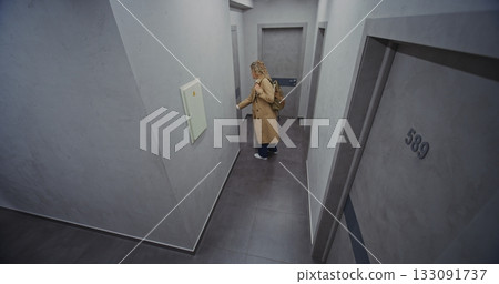 Stylish Female Traveler in Light Trench Coat Walks Down Hotel Corridor 133091737