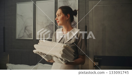 Smiling Woman in Neat Professional Uniform Holds Folded Stack of Fresh White Towels in Hotel Room Smiling Woman in Neat Professional Uniform Holds Folded Stack of Fresh White Towels in Hotel Room 133091745