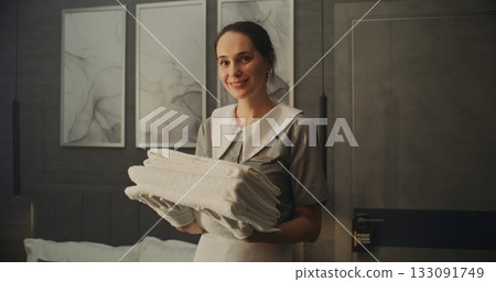 Smiling Woman in Neat Professional Uniform Holds Folded Stack of Fresh White Towels in Hotel Room 133091749