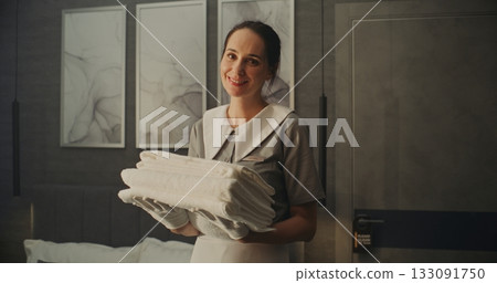 Smiling Woman in Neat Professional Uniform Holds Folded Stack of Fresh White Towels in Hotel Room Smiling Woman in Neat Professional Uniform Holds Folded Stack of Fresh White Towels in Hotel Room 133091750