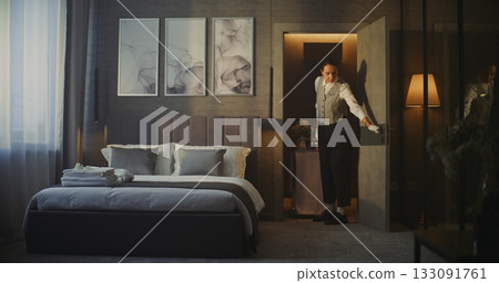 Young Hotel Employee in Uniform Brings Room Service Trolley Into Modern Room in Luxury Hotel 133091761