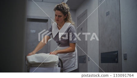 Female Hotel Housekeeper in Neat Uniform Stands Beside Cleaning Trolley, Carefully Folding Towels 133091764