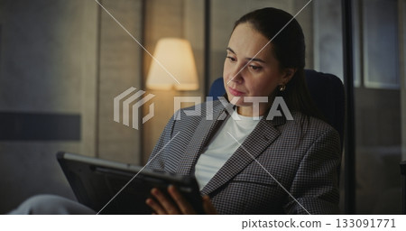 Woman With Long Dark Hair Sits Comfortably in Modern Hotel Armchair, Holding Tablet on Lap 133091771