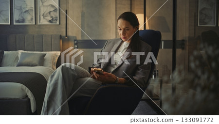 Stylish Woman With Long Dark Hair Sits in Soft Armchair in Hotel Room With Phone in Hands Stylish Woman With Long Dark Hair Sits in Soft Armchair in Hotel Room With Phone in Hands 133091772