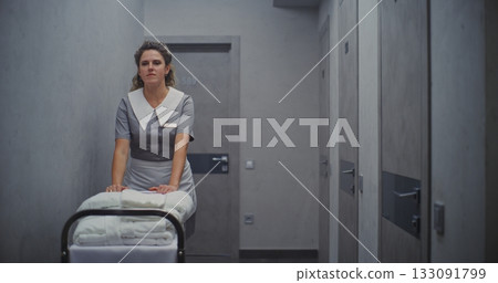Female Maid in Professional Uniform Moves Cleaning Trolley Down Hallway, Carrying Neatly Towels 133091799