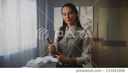 Smiling Young Female Hotel Service Professional in Uniform Holds Tablet in Modern Luxury Hotel Room 133091806