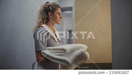 Curly Haired Hotel Housekeeper in Uniform Pushes Cleaning Trolley Through Bright Hallway 133091807