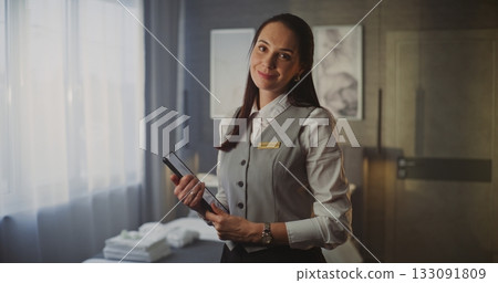 Smiling Young Female Hotel Service Professional in Uniform Holds Tablet in Modern Luxury Hotel Room 133091809