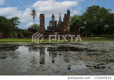A serene Buddha image stands amid brick ruins beside a lotus-filled pond, creating a tranquil historical sanctuary at Sukhothai Historical Park 133092610