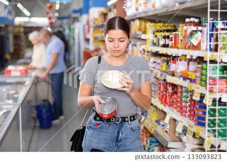 Young woman purchaser choosing tinned fish in big supermarket 133093823