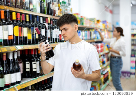 in supermarket young man carefully chooses wine in supermarket young man carefully chooses wine 133093839
