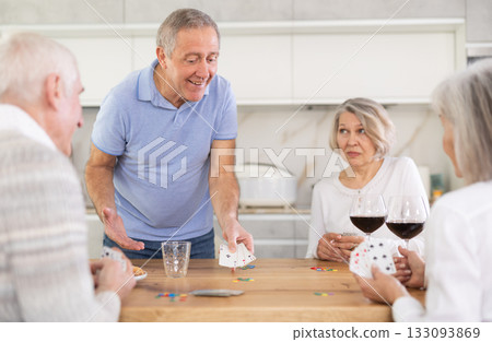 Cheerful friendly company of men and women sitting at kitchen table and enjoying the poker game. Friends play card game at home. Evening entertainment concept 133093869