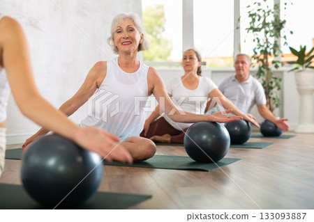 Elderly woman posing with soft ball at group class in modern yoga studio Elderly woman posing with soft ball at group class in modern yoga studio 133093887